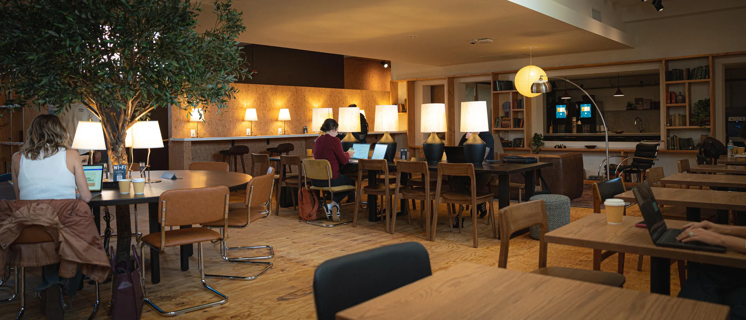 Inside The Work Club: members working at wooden tables under warm lamps, a tree in the middle of the room and the coffee bar at the back.